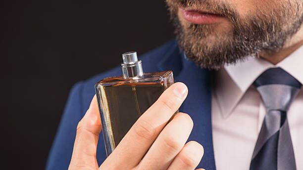Homem de barba e e terno azul e segurando um frasco de perfume