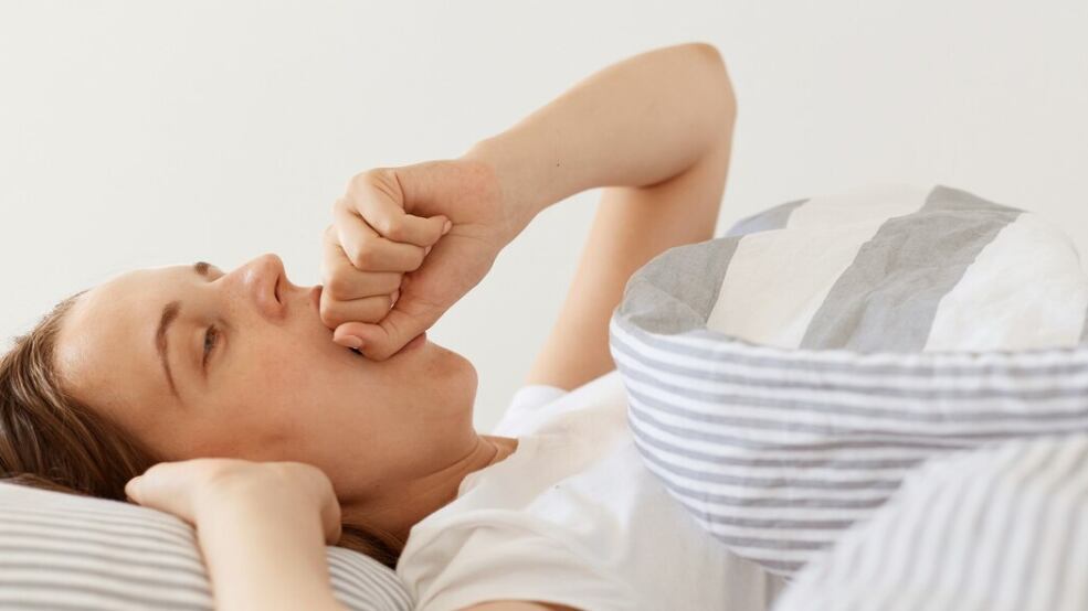 Mujer soñolienta con cabello oscuro con camiseta blanca casual acostada debajo de una manta y bostezando, tapándose la boca con el puño, simplemente despierta, disfrutando temprano en la mañana.