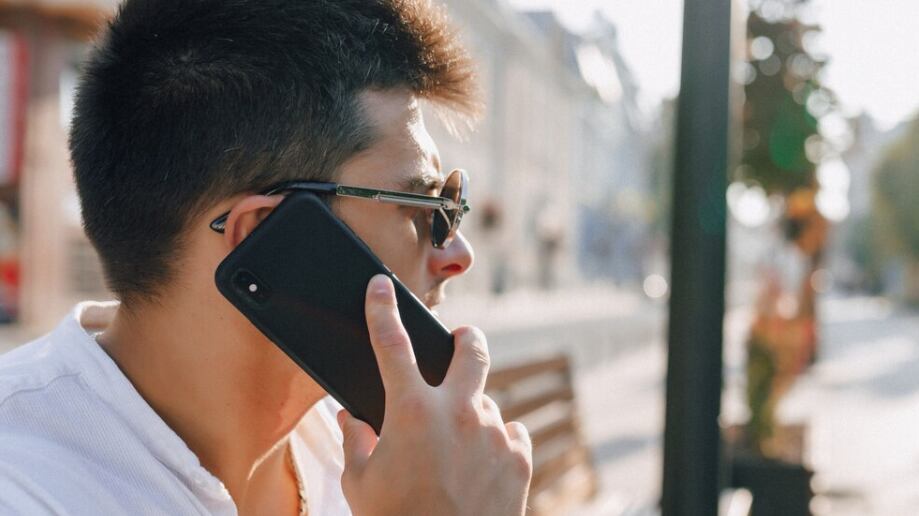 Chico con estilo joven en camisa con teléfono en banco en día soleado al aire libre.